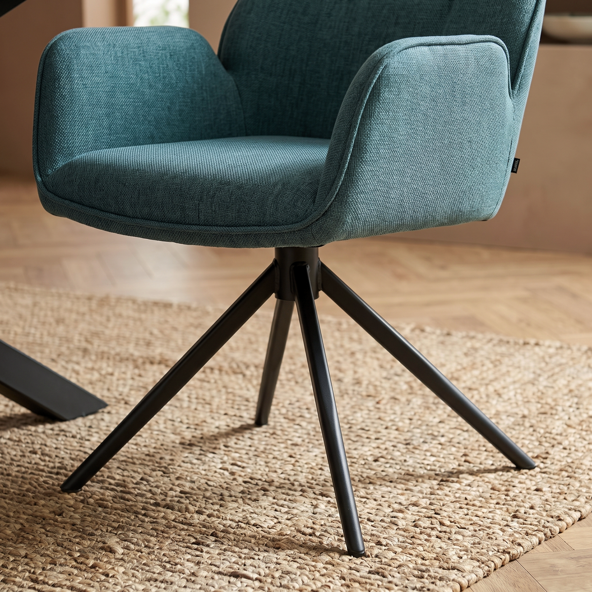Close up of a Benicio teal swivel dining chair, showing the woven fabric seat and arm and the black four pronged metal swivel base
