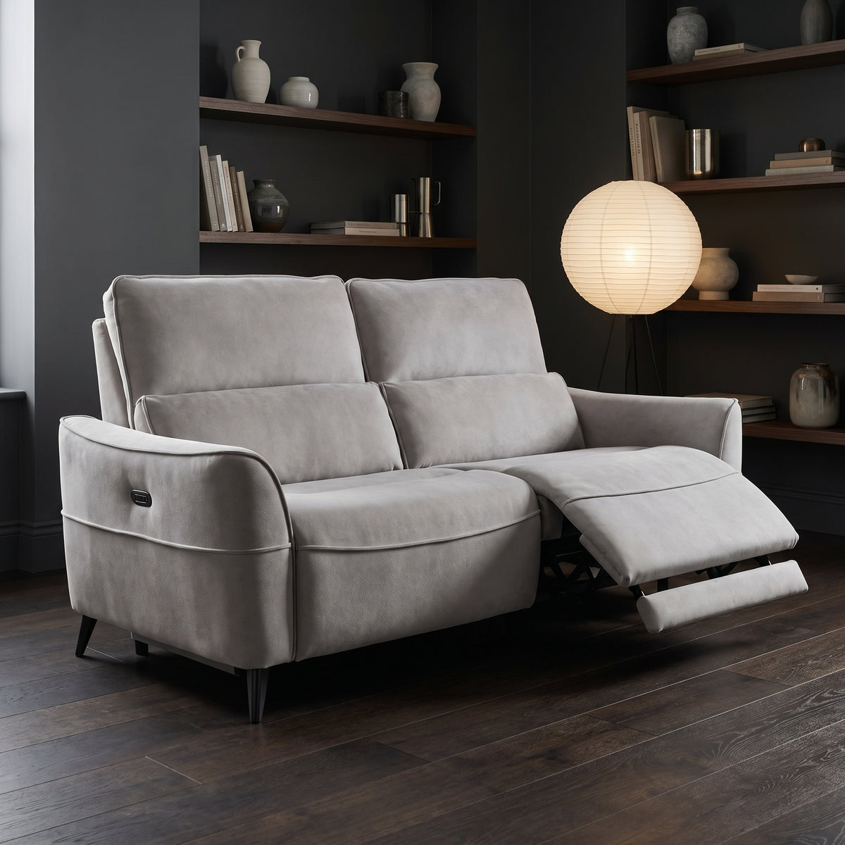 Dune electric recliner in dove grey with one footrest extended, in a dark walled room with a paper lantern light