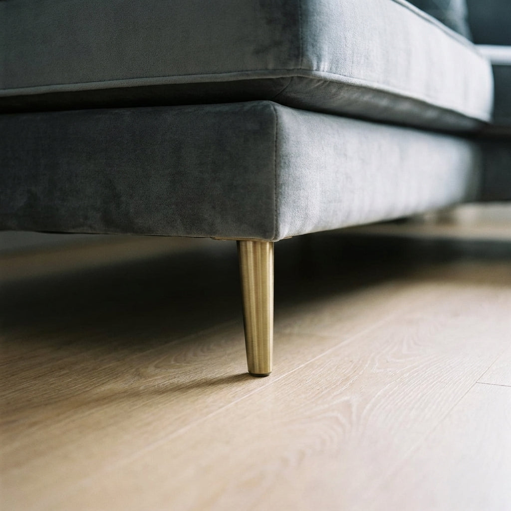 Close-up of a modern grey velvet sofa with elegant gold tapered legs on light wood flooring in a contemporary living room.