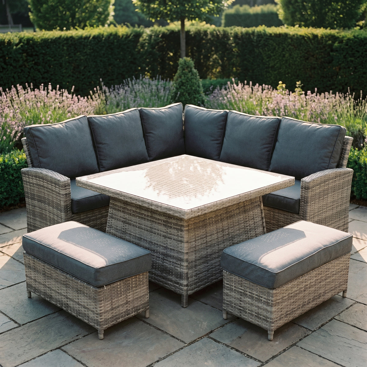 Maldives grey rattan corner garden set with glass top table and two stools, on a stone patio with lavender borders